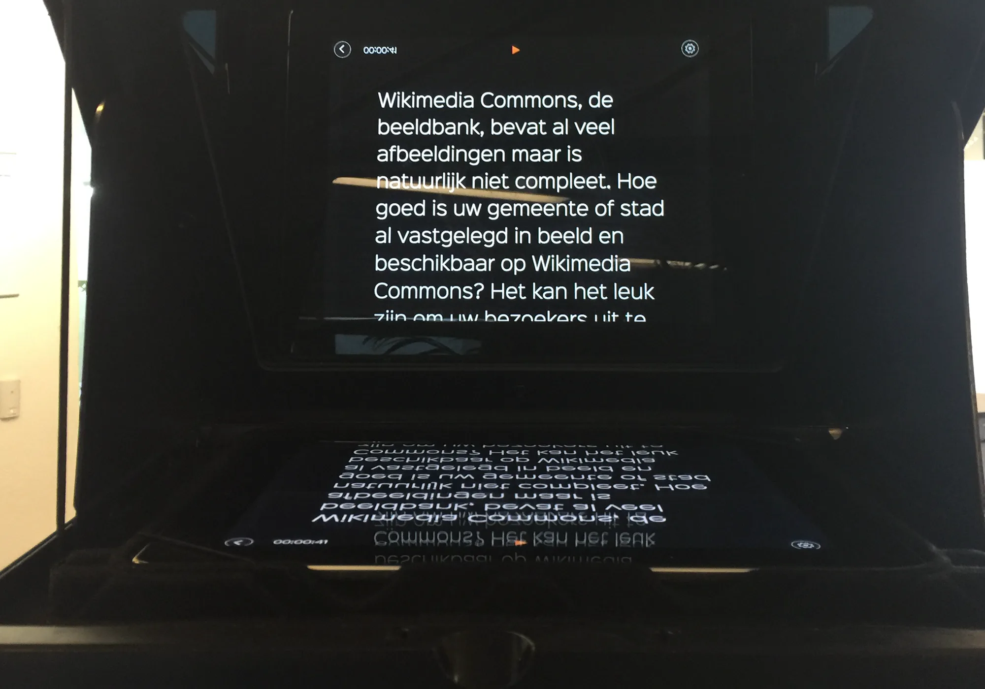 A tablet teleprompter seen from the presenter's side. White script text reads normally on the angled glass, and on the tablet lying below it the same lines appear upside down and mirrored