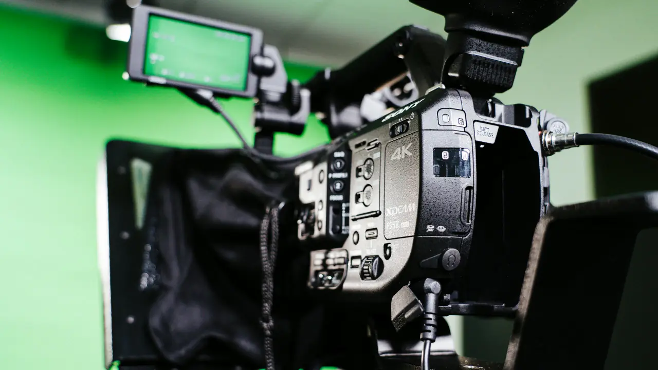 A video camera mounted behind a teleprompter hood in a green-screen studio, seen from behind the camera