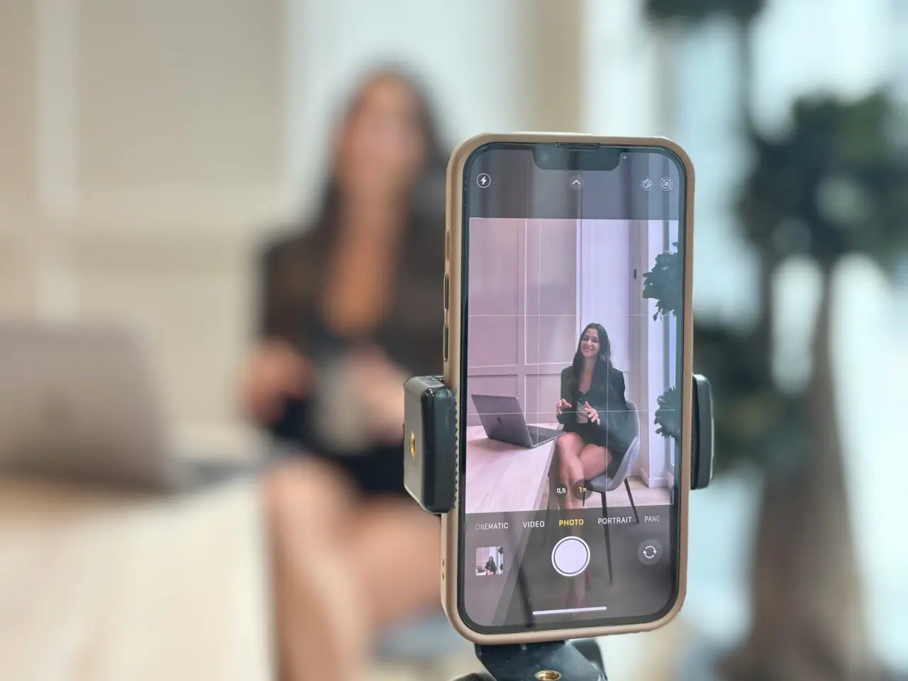 An iPhone on a small tripod in the foreground, its camera framing a woman sitting at a desk with a laptop, who is out of focus behind it