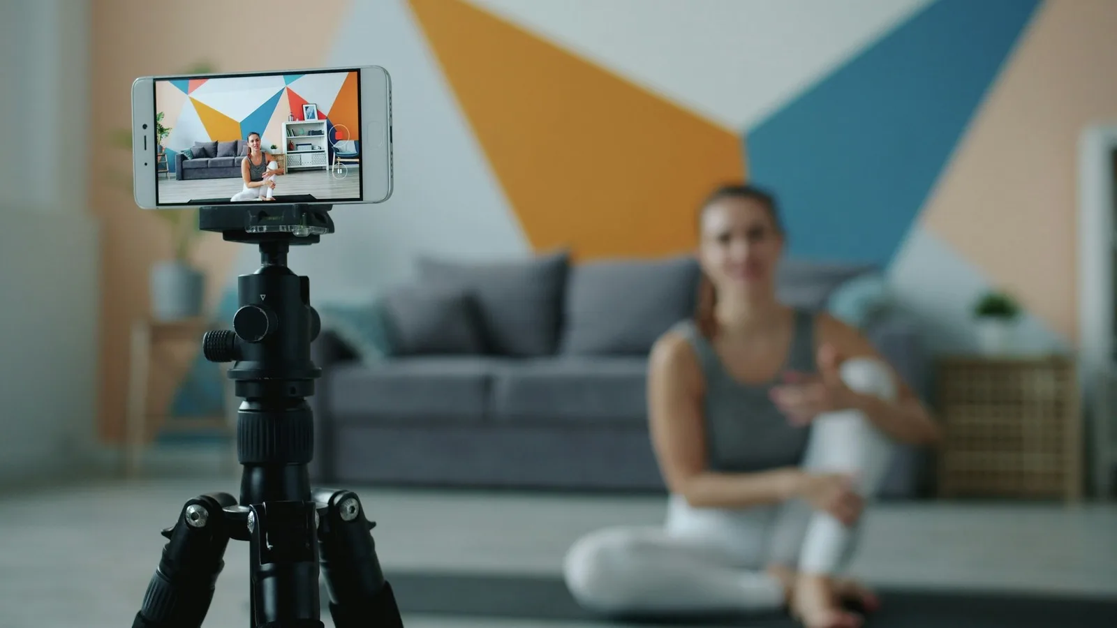 An iPhone on a tripod, turned sideways, filming a woman sitting on a mat in a bright living room