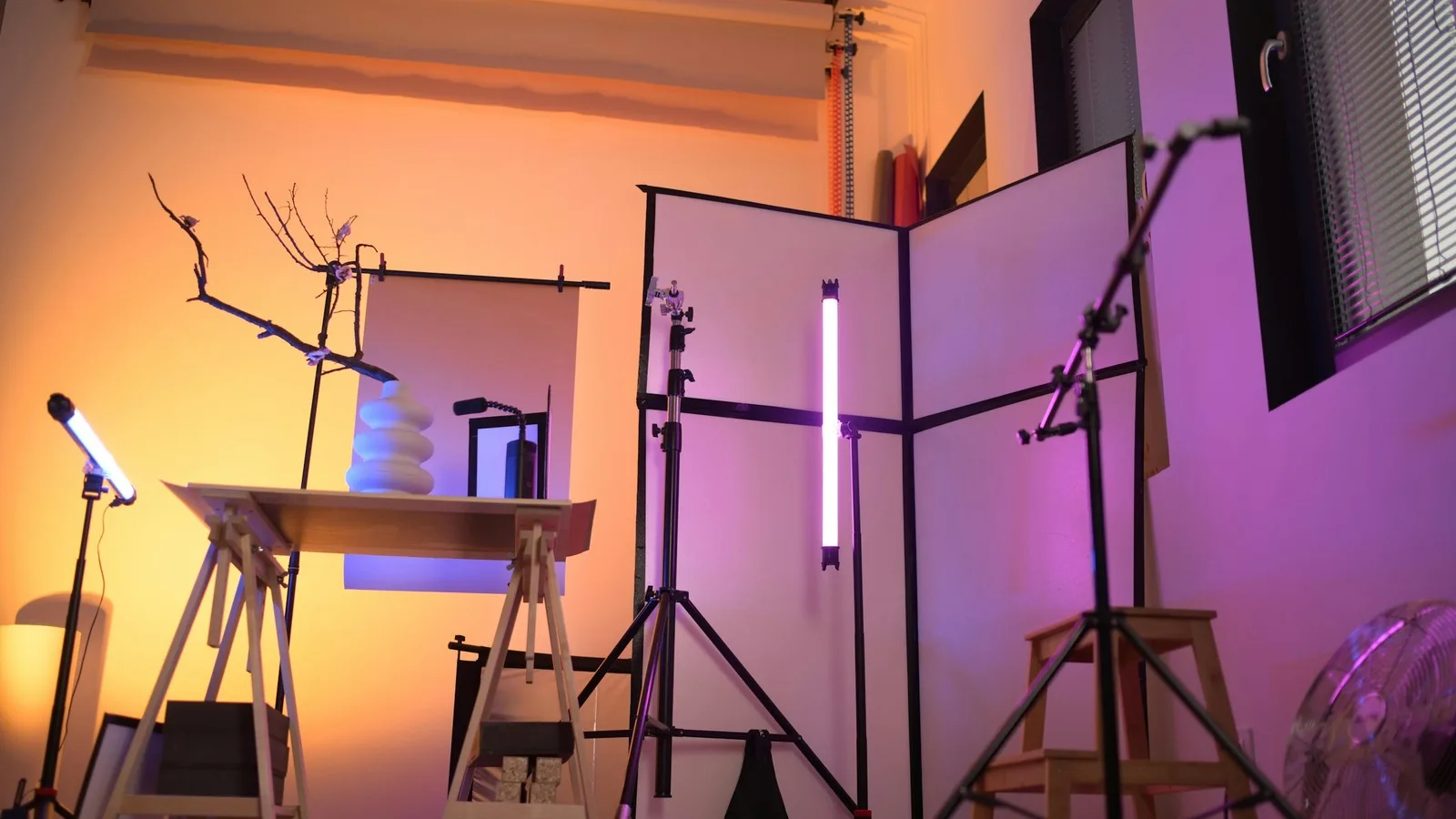 A small home studio lit with warm and violet LED tube lights, with stands, a white backdrop and a desk