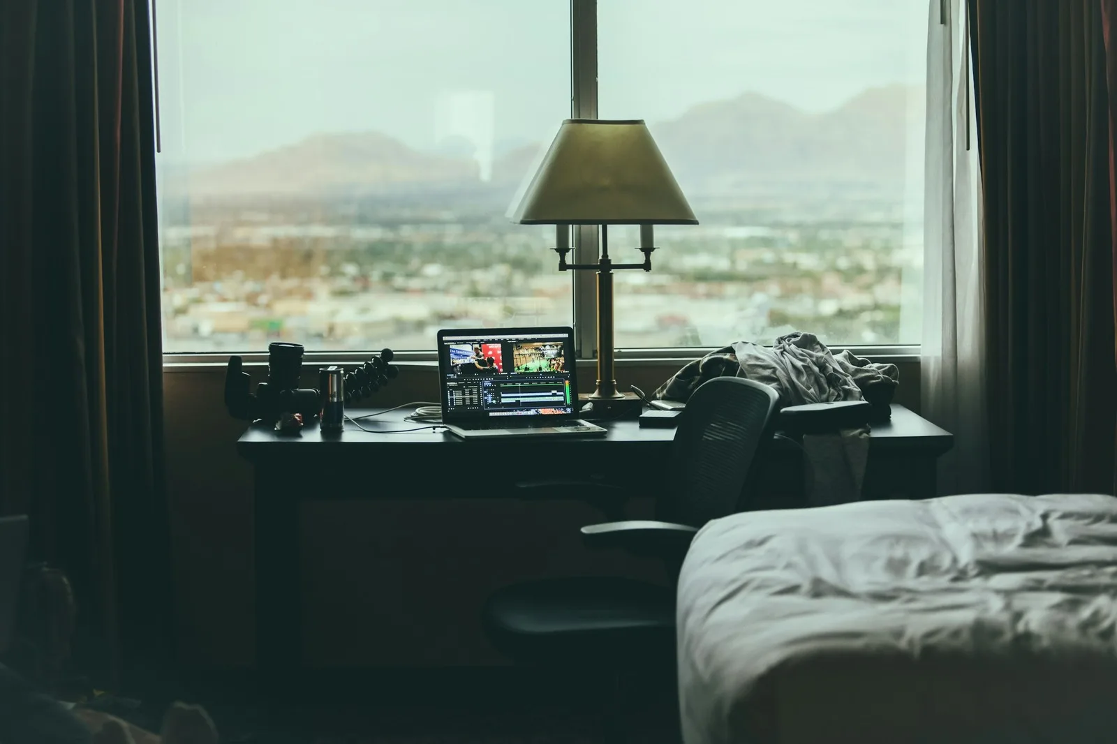A desk set right in front of a large window with a view over a city, with a laptop, a lamp and a chair facing the glass