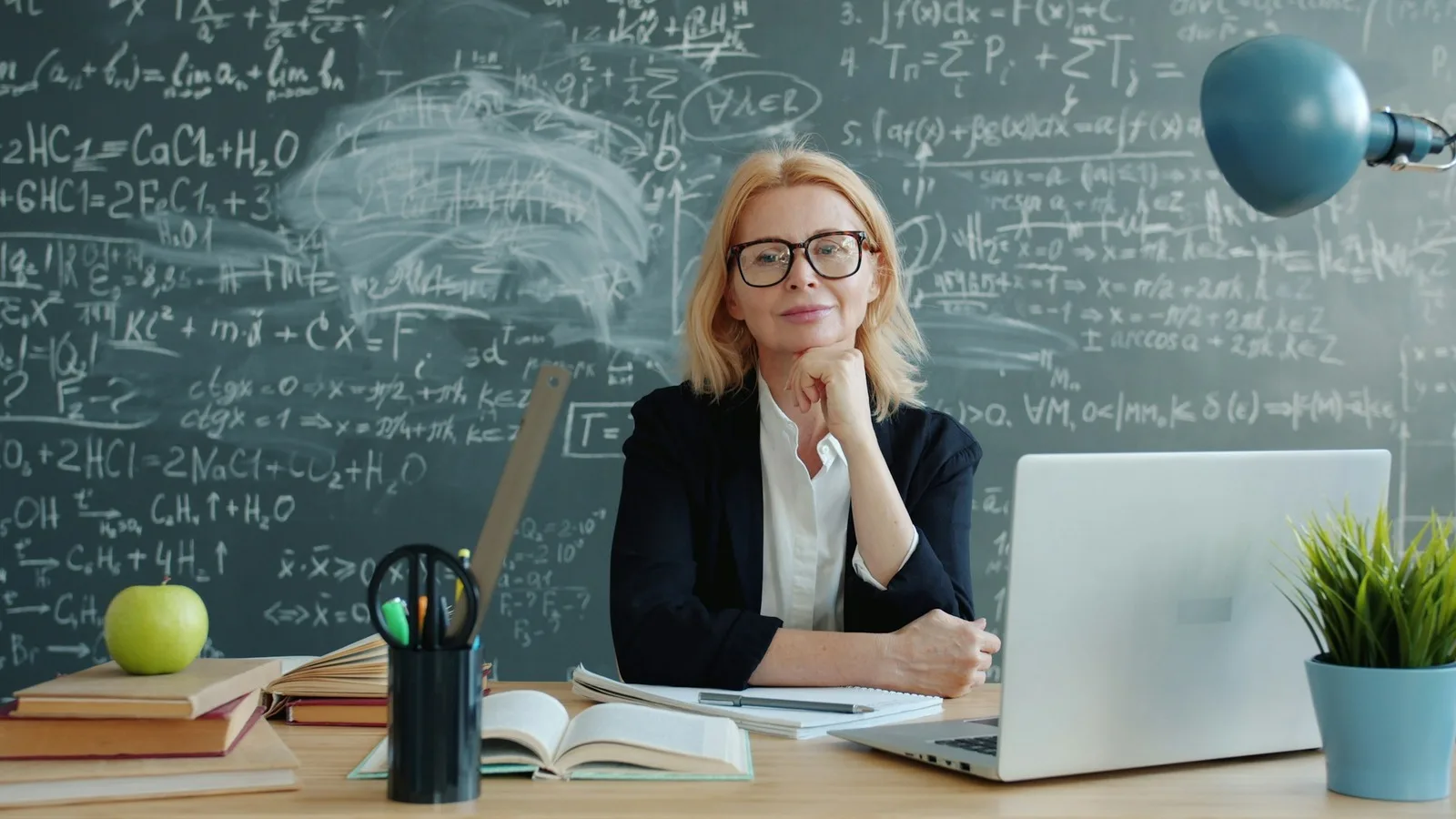 A teacher in glasses sitting at a desk with a laptop, in front of a chalkboard covered in equations