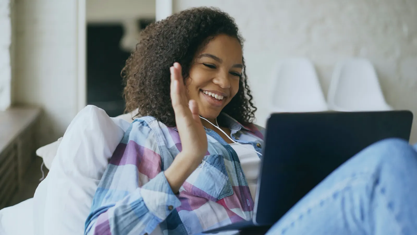 A woman smiling and waving at her laptop's camera from a sofa