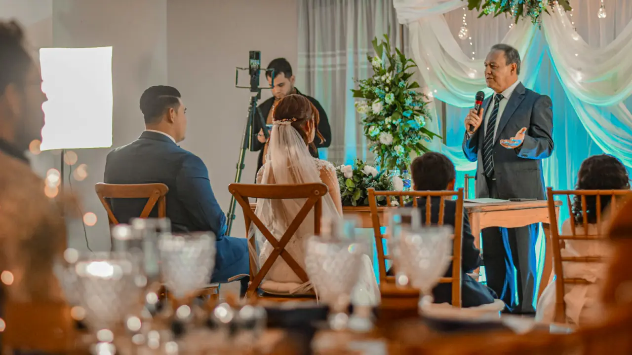 A guest in a suit giving a speech with a microphone at a wedding reception, while the bride and groom listen from their chairs